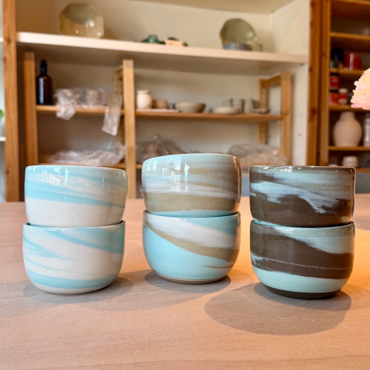 Three pairs of stacked ceramic cups with a marbled swirl pattern in light blue, beige, and brown, arranged on a wooden table with a pottery shop in the background.