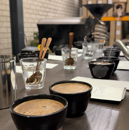 Two black ceramic coffee cups filled with espresso on a wooden table in a cafe setting, with glass cups, wooden spoons, and a notebook nearby.