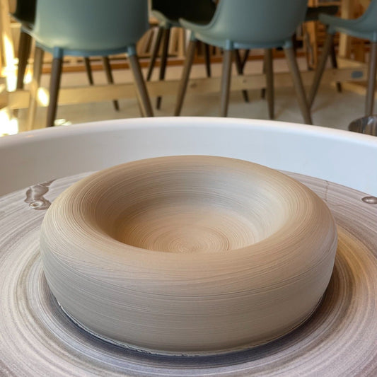 A handmade, unglazed ceramic bowl or planter on a pottery wheel, with a blurred pottery studio background featuring shelves, chairs, and a table.
